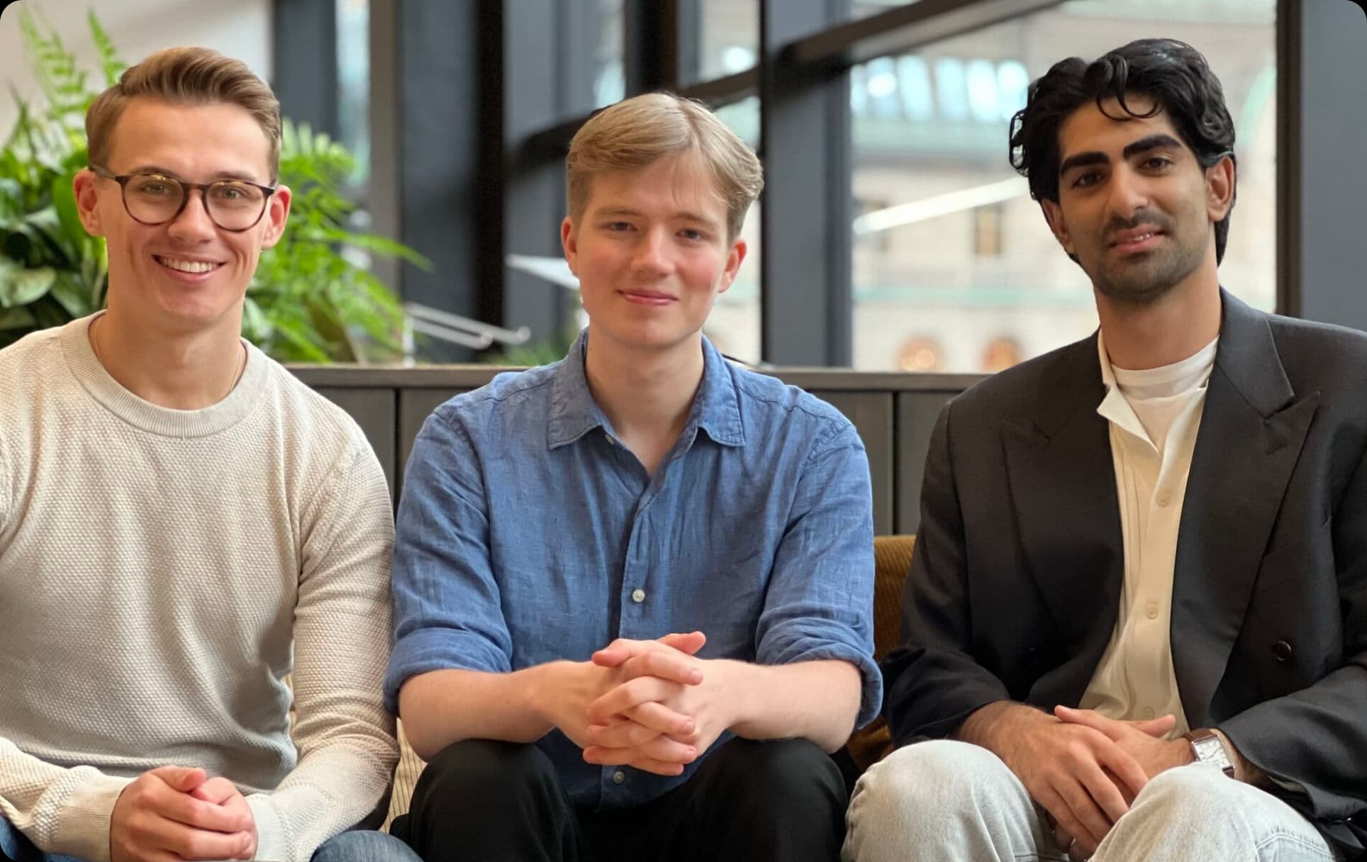 Three individuals sit closely together on a sofa, dressed in casual and smart attire, surrounded by greenery in a modern indoor space.